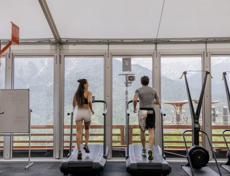Two people running on treadmills in a gym with scenic mountain views.