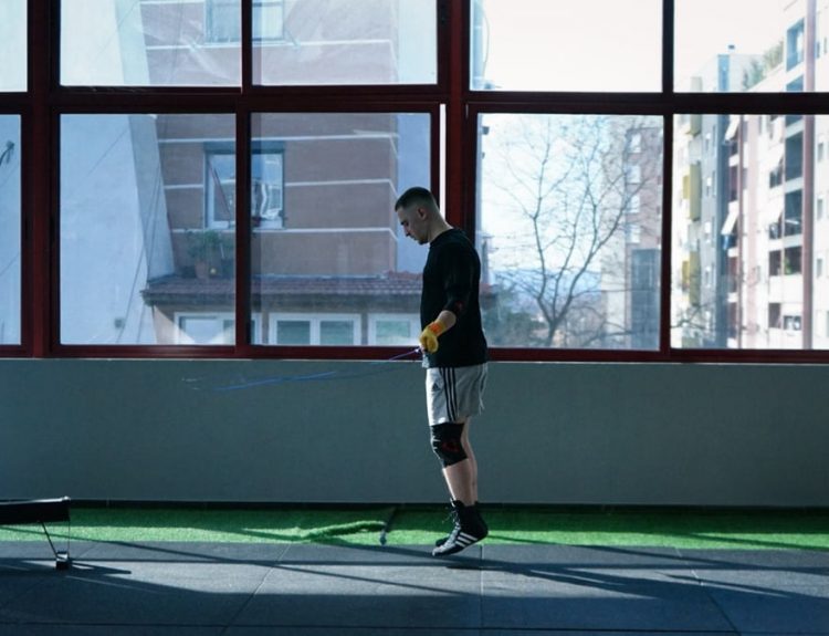 Young man exercising with a skipping rope in an urban gym with large windows and city view.